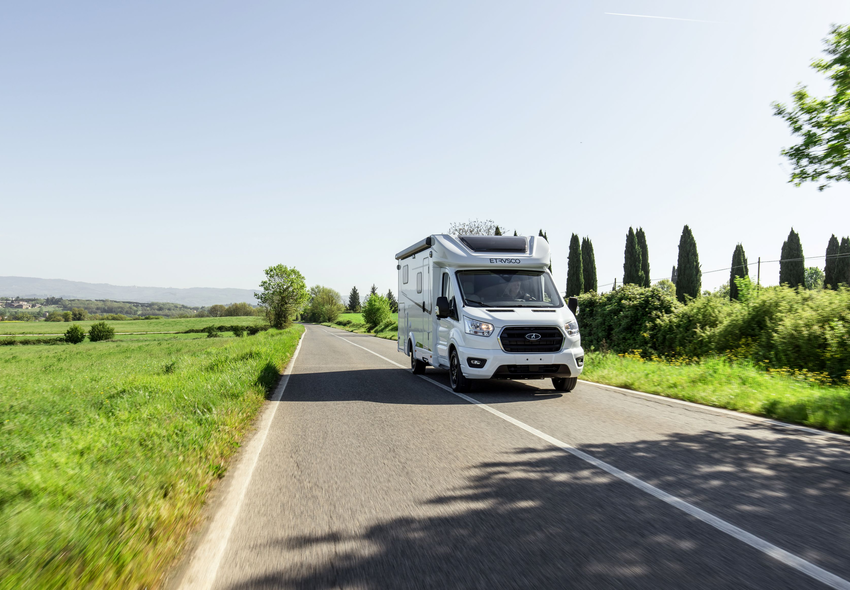 Ein schmaler Teilintegrierter Etrusco Van Ford Wohnmobil fährt auf einer Landstraße vor einem Hintergrund mit Bergen und rötlichem Himmel.