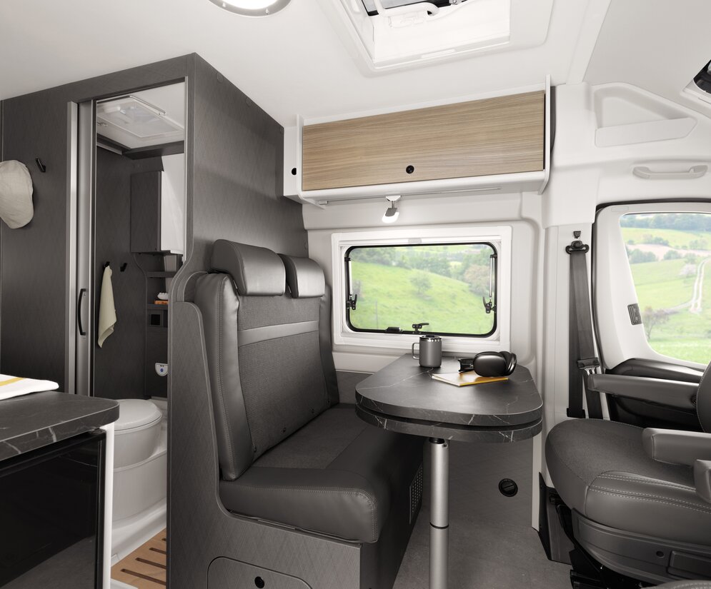 Bestuurdersgebied van een Etrusco Campervan Fiat Plus met draaibare stoelen, panoramavenster en koffietafel, omgeven door groen landschap.