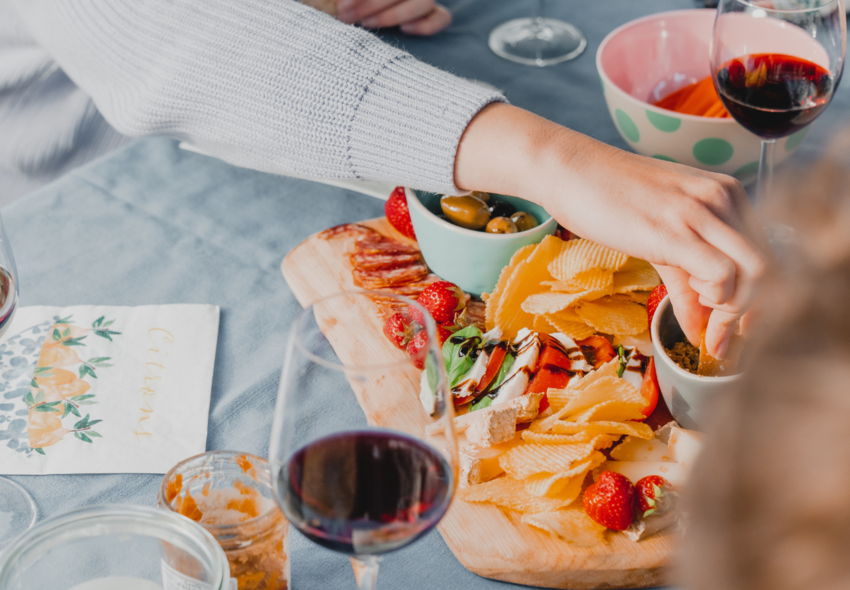 Campingmaaltijd met snacks, olijven, aardbeien, kaas, rode wijn en een hand op de tafel, ideaal voor samenzijn tijdens het kamperen.