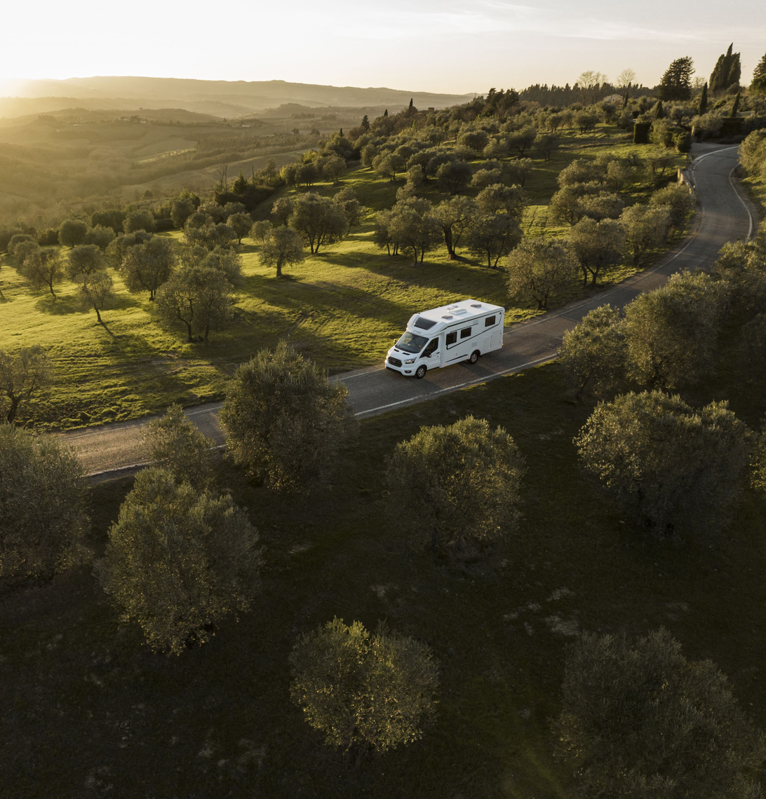 En delintegreret Etrusco autocamper på Ford-basis kører gennem et malerisk landskab med oliventræer i den gyldne solnedgang.