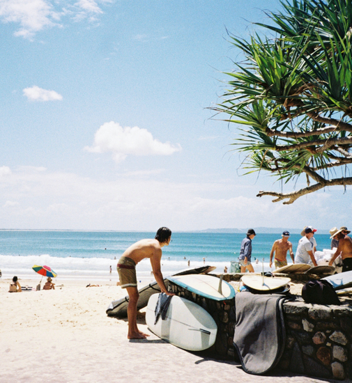 Sonniger Strand mit Surfbrettern und einem Mann im Vordergrund – perfekte Atmosphäre für Campingausflüge und Wassersport.