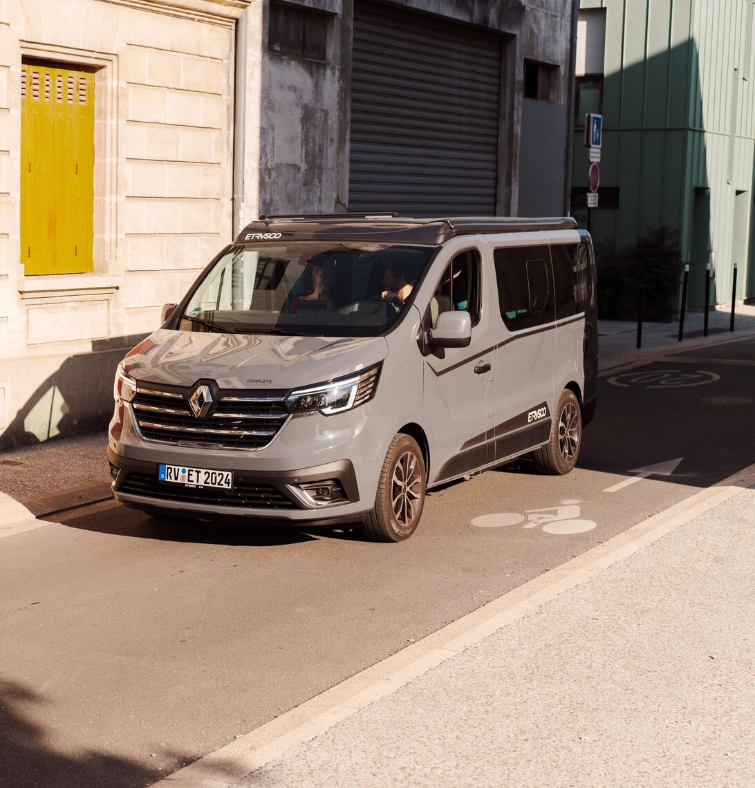Etrusco UC 5.0 XR Urban Camper grigio su base Renault percorre una strada soleggiata attraverso una città mediterranea.