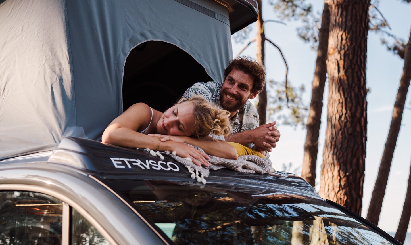  Un uomo e una donna si rilassano sul tetto sollevato di un Etrusco UC 5.0 XR Urban Camper circondati da alberi e cielo blu.
